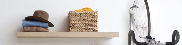 A bicycle hangs from a wall mount hook next  to a shelf with some clothing, a hat and a basket on it.