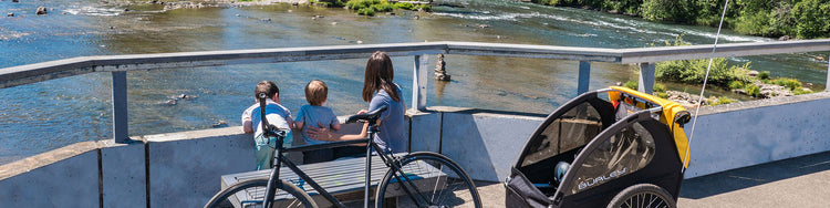 Amom and her two children stop to look at a river int he background while her bike and Burley child trailer are in the foreground.