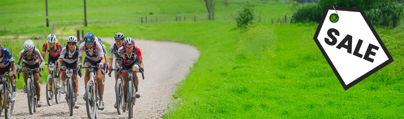 A pack of gravel riders in helmets, sunglasses and cycling clothing ride down a gravel road in the countryside. A black and white "sale" graphic overlays the image.