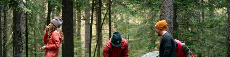 Three campers in the woods are wearing hats of assorted colors.