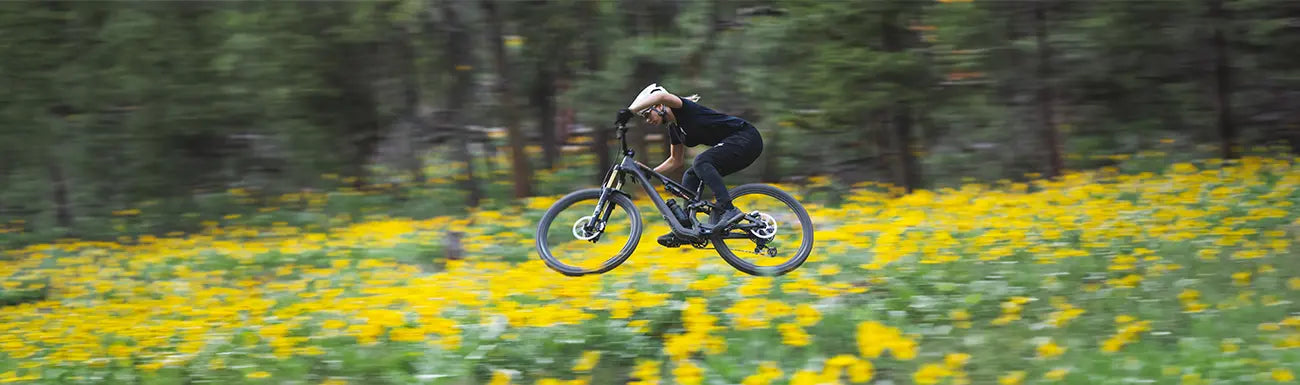 A woman rides a Specialized Mountain bike through a wooded area with yellow flowers in the foreground.