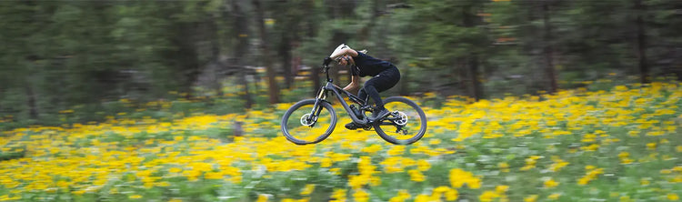 A woman rides a Specialized Mountain bike through a wooded area with yellow flowers in the foreground.