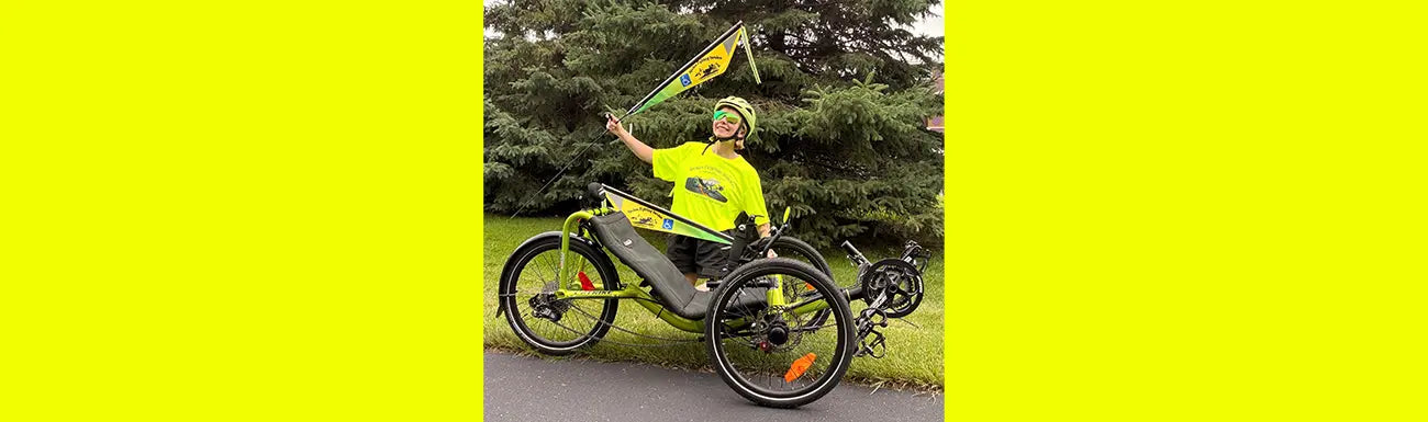 A person wearing a bright neon Spokes Fighting Strokes t-shirt, matching helmet and green sunglasses sits on a lime green recumbent trike with evergreen trees in the background. The trike has a matching yellow-green Spokes Fighting Strokes flag.