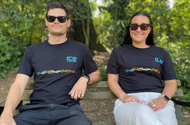 Front view of a man and woman wearing matching black t-shirt with a light blue ICE logo above the left chest and five recumbent trikes in blue, orange, white, green and red extending across the chest below the logo. 
