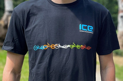 Front view of person wearing a black t-shirt with a light blue ICE logo above the left chest and five recumbent trikes in blue, orange, white, green and red extending across the chest below the logo. 