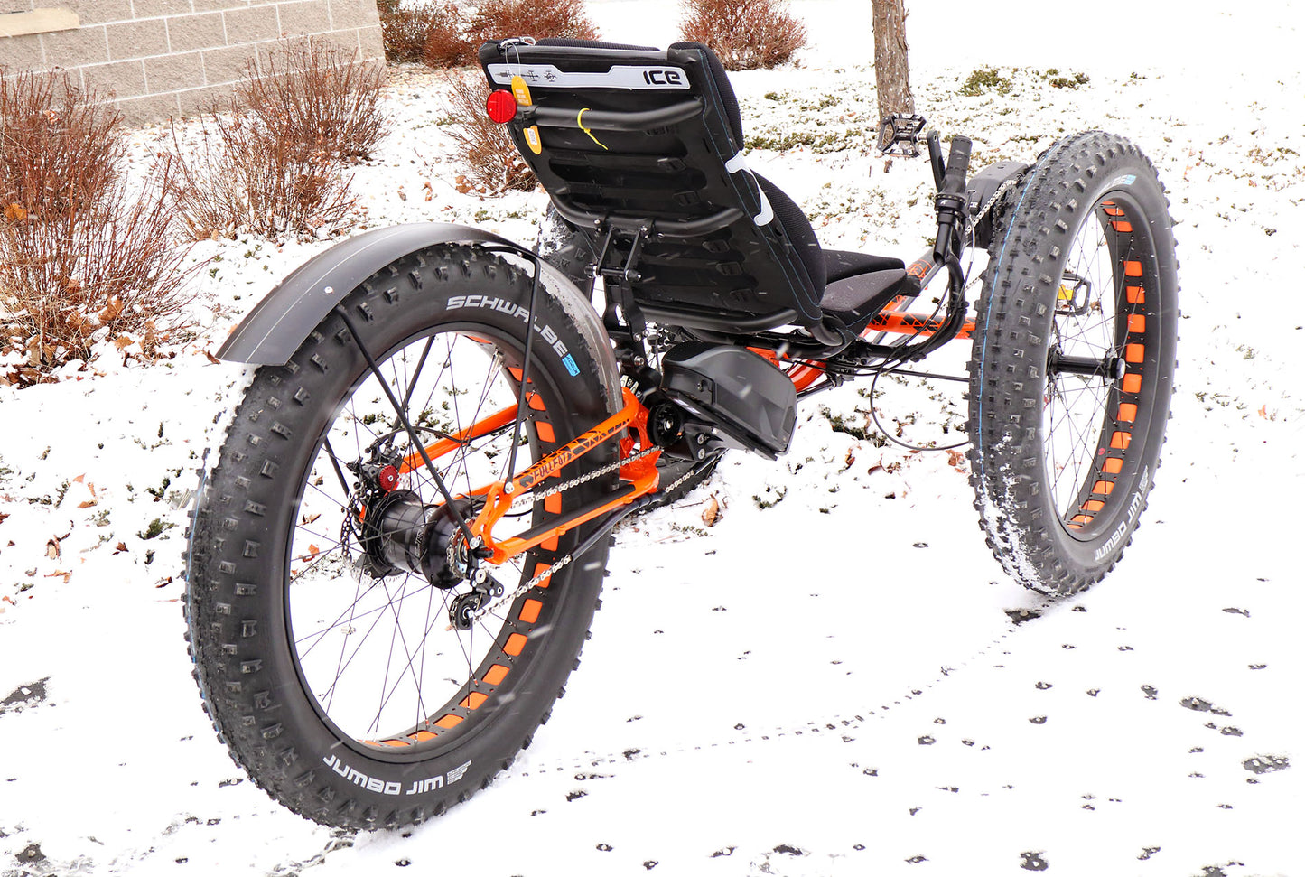 Right back angle view of an ICE full fat recumbent trike with black seat, tires, battery, and rear fender and orange and black frame. 
