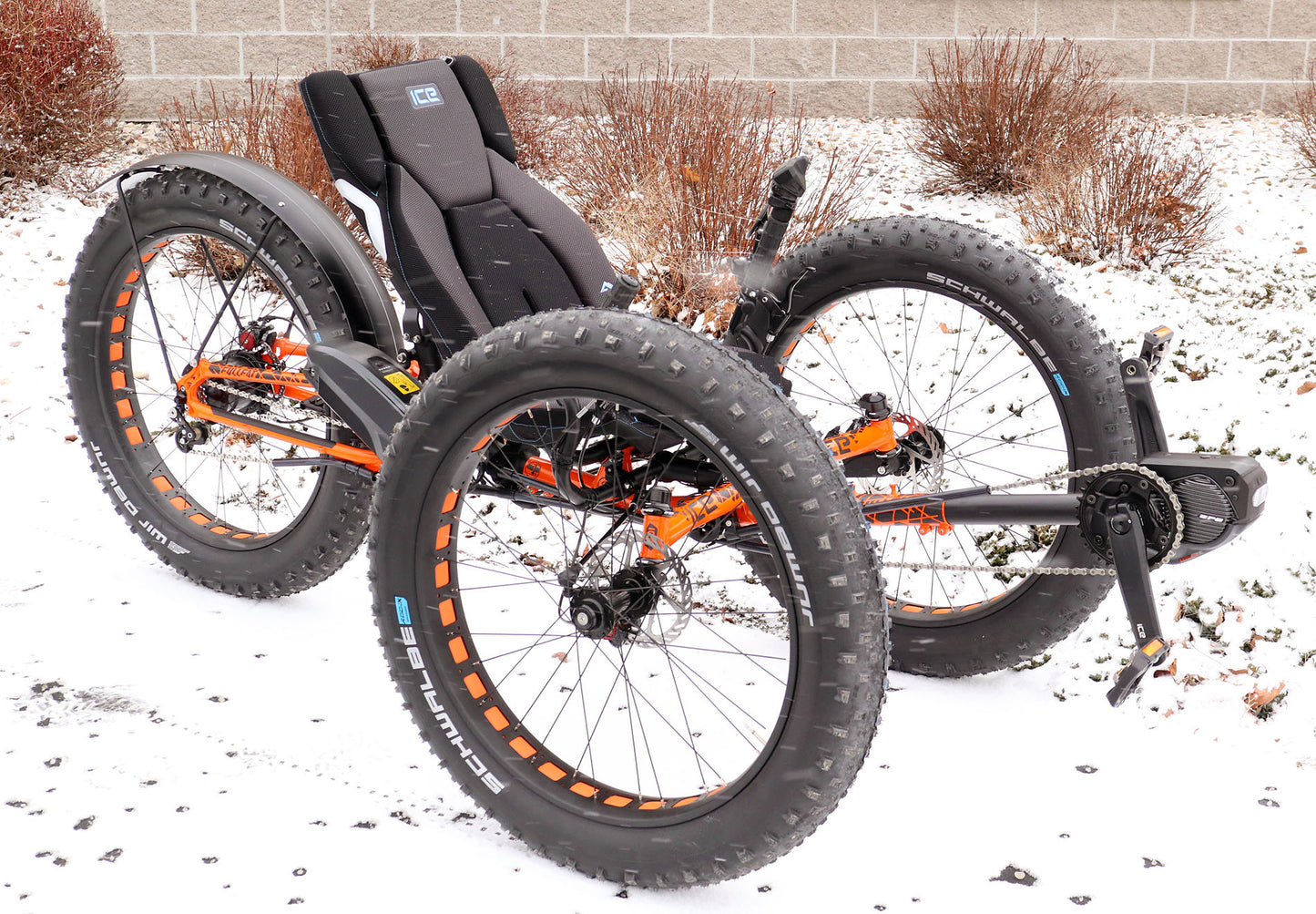 Front right side view of an orange ICE full fat recumbent trike in the snow. 