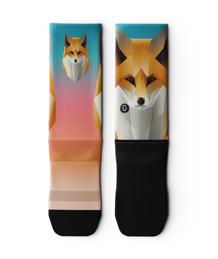 Multicolored pair of socks laying flat, with geometric sock head. Background of sock is teal blue at the top fading to light orange near toes. Sole of socks is black