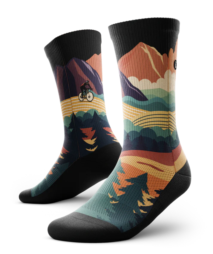 Pair of multicolored socks with black soles depicting mountain forest scene with person riding bike on a trail.