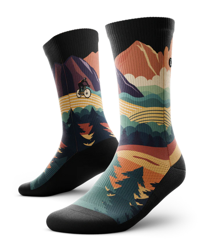 Pair of multicolored socks with black soles depicting mountain forest scene with person riding bike on a trail.