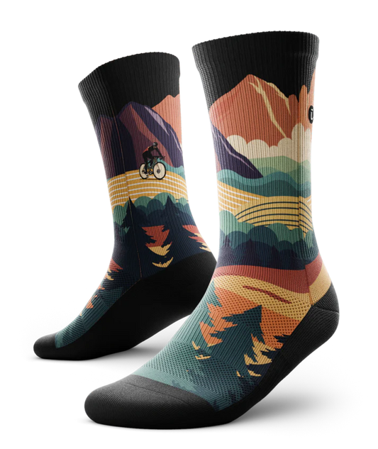 Pair of multicolored socks with black soles depicting mountain forest scene with person riding bike on a trail.