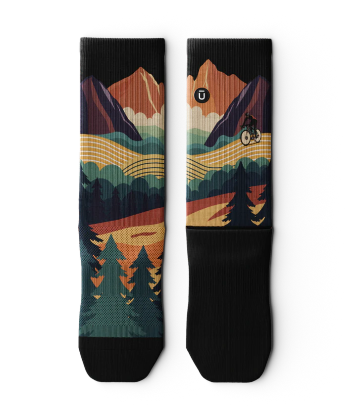 Pair of multicolored socks laying flat  with black soles depicting mountain forest scene with person riding bike on a trail.