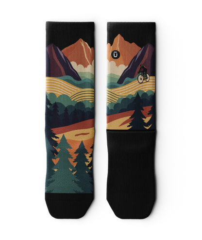 Pair of multicolored socks laying flat  with black soles depicting mountain forest scene with person riding bike on a trail.