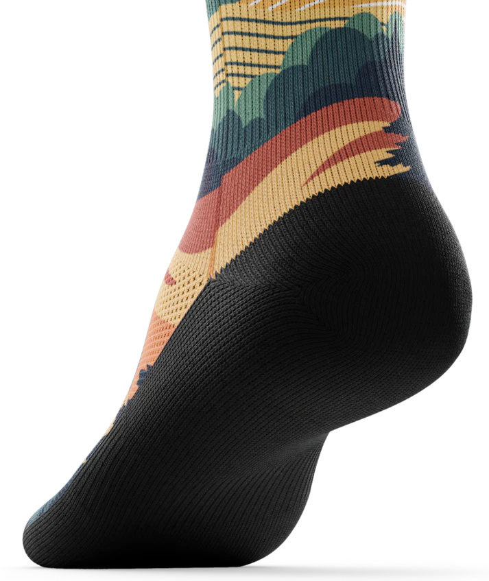 Bottom black sole of a multicolored sock depicting mountain forest scene with person riding bike on a trail.