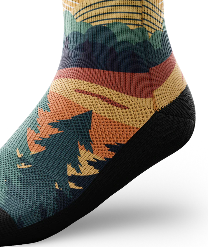 Ankle view of a multicolored sock depicting mountain forest scene with person riding bike on a trail.