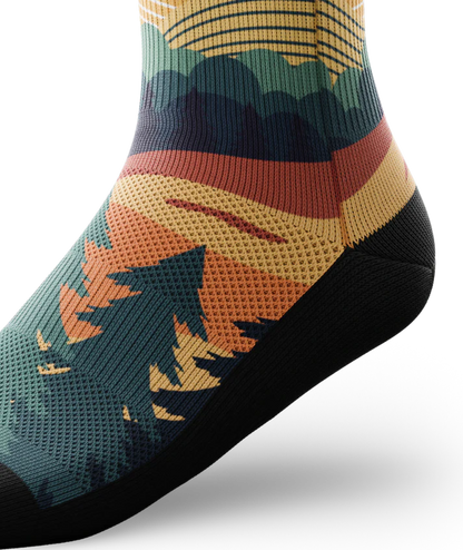 Ankle view of a multicolored sock depicting mountain forest scene with person riding bike on a trail.