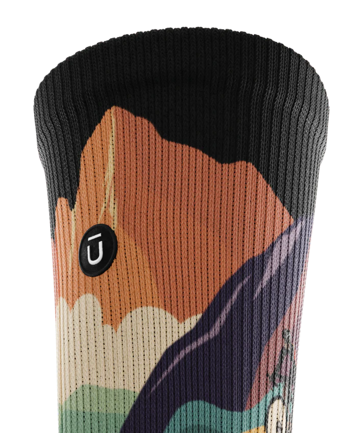 Top cuff view of a multicolored sock depicting mountain forest scene with person riding bike on a trail.  Small black circular patch with Outway logo stitched on top third of cuff. 
