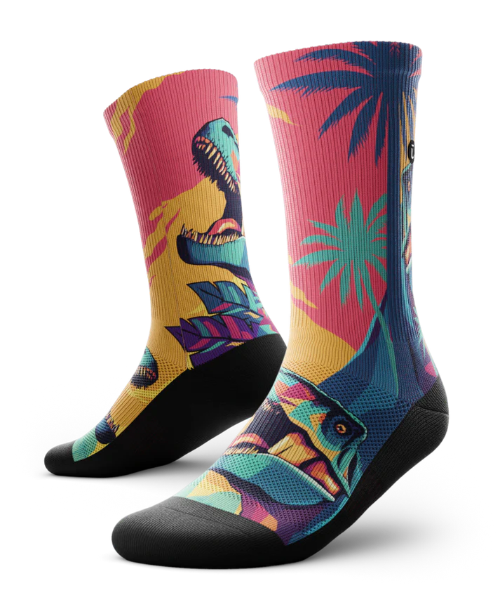 Multicolored  pair of socks with T-rex dinosaurs and palm trees on main part of sock, black soles. 