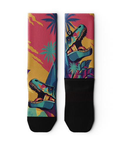 Multicolored  pair of socks laying flat with T-rex dinosaurs and palm trees on main part of sock, black soles. 