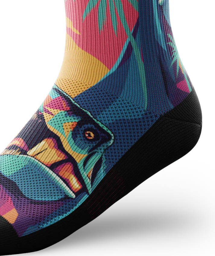 Side ankle view of Multicolored sock with T-rex dinosaurs and palm trees on main part of sock, black soles. 