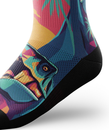 Side ankle view of Multicolored sock with T-rex dinosaurs and palm trees on main part of sock, black soles. 