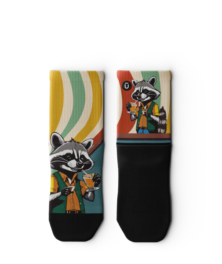 Multicolored pair of socks laying flat with racoon wearing brown button down shirt and green vest drinking cups of coffee. Swirled cuff of green, cream, yellow and red. Black soles
