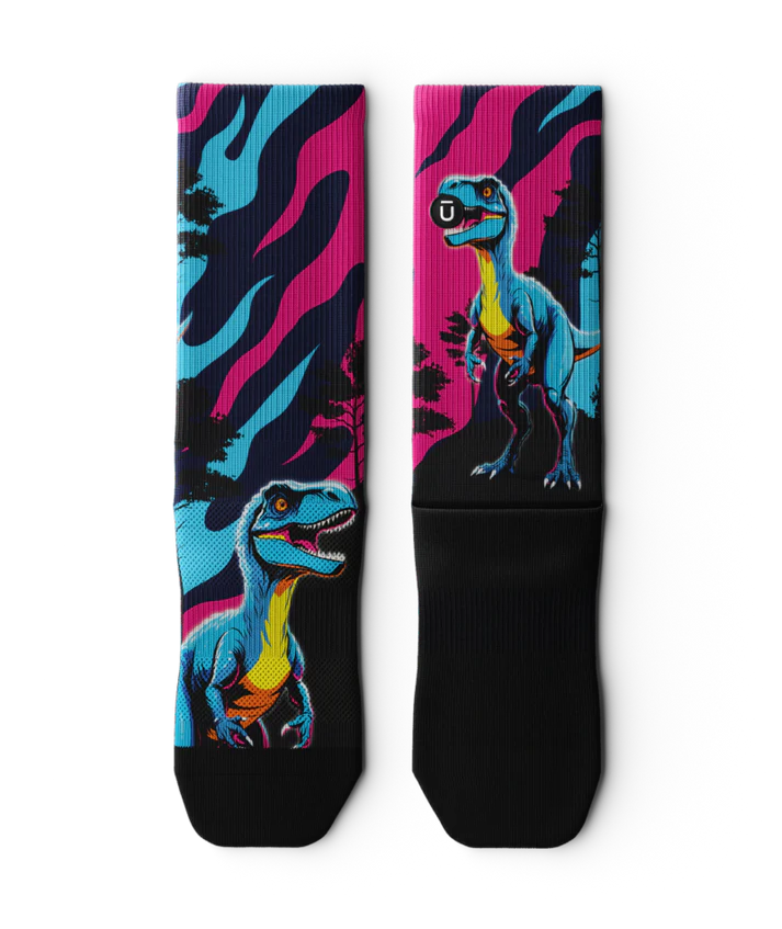 Multicolored pink, blue and black pair of socks  laying flat with velociraptors, black trees and black soles. 