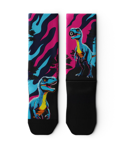 Multicolored pink, blue and black pair of socks  laying flat with velociraptors, black trees and black soles. 