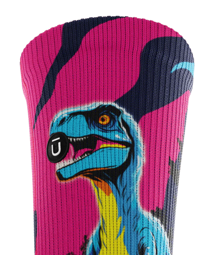 Top cuff of multicolored pink, blue and black pair of socks with velociraptors, and black trees. Small black circular patch with Outway logo on top third of cuff. 