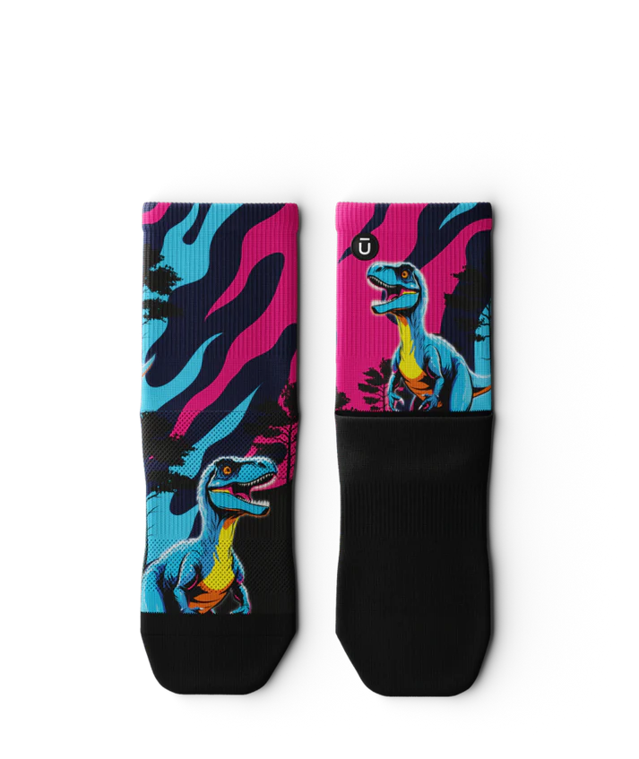 Multicolored pink, blue and black pair of socks  laying flat with velociraptors, black trees and black soles. 