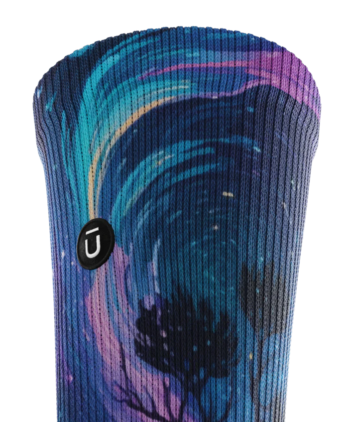 Top cuff view of multicolored sock with colorful northern lights in the ski over a mountain lake with trees silhouetted.  Small black circular patch with Outway logo on op third of cuff. 
