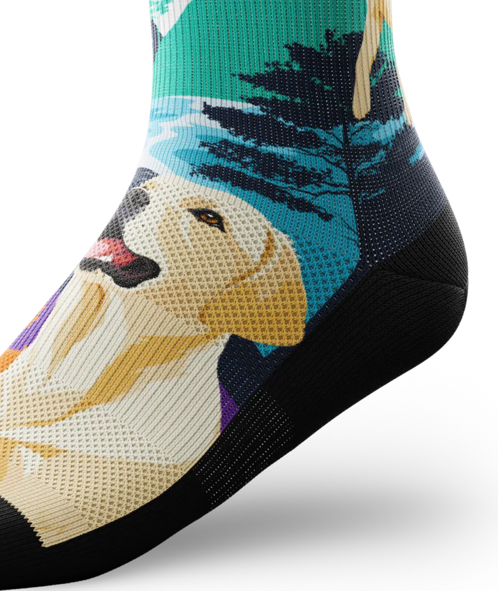 Ankle view of multicolored sock with yellow labs chasing a tennis ball on a string, with a Asian inspired background of sun, trees, mountain and stream. 