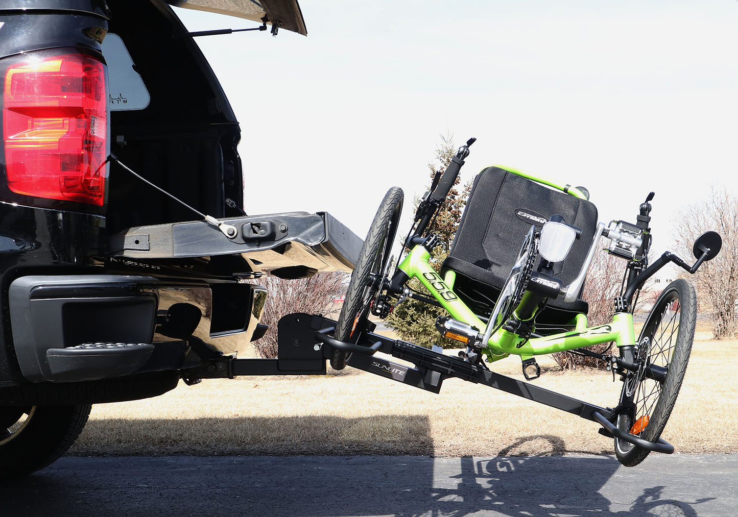 Tilted down view of the Sunlite HRT-100 Trike Auto Rack for 2-inch Receivers mounted on the hitch on a pickup truck to allow access to the tailgate with a green Catrike on the rack in an outdoor setting.