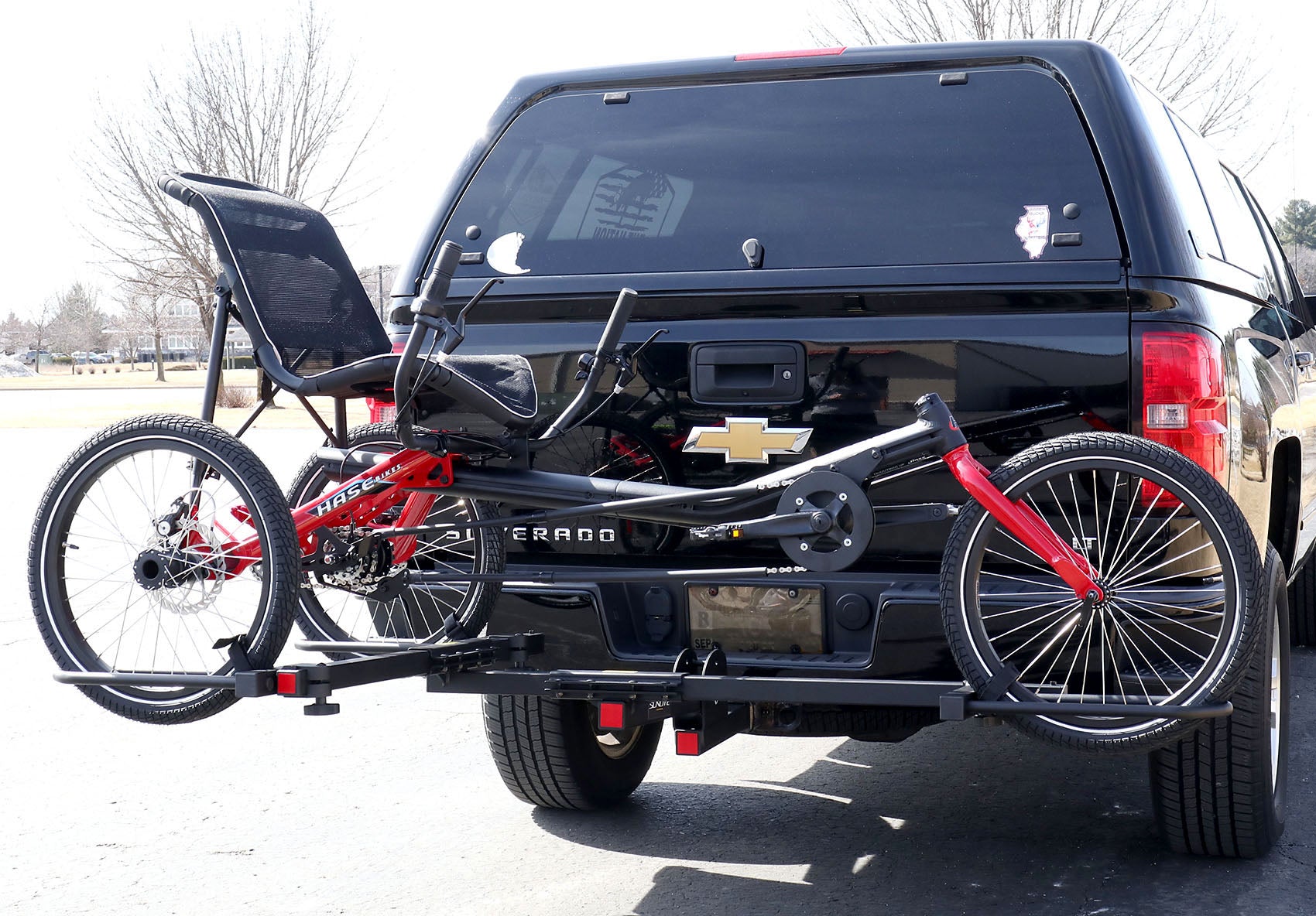 Angled rear view of the Sunlite HRT-100 Trike Auto Rack for 2-inch Receivers mounted on the hitch on a pickup truck  with a red Hase Trigo on the rack in an outdoor setting.