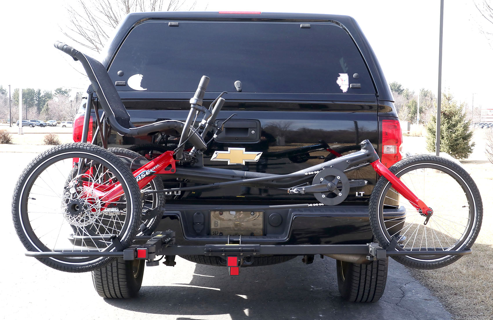 Rear view of the Sunlite HRT-100 Trike Auto Rack for 2-inch Receivers mounted on the hitch on a pickup truck  with a red Hase Trigo on the rack in an outdoor setting.