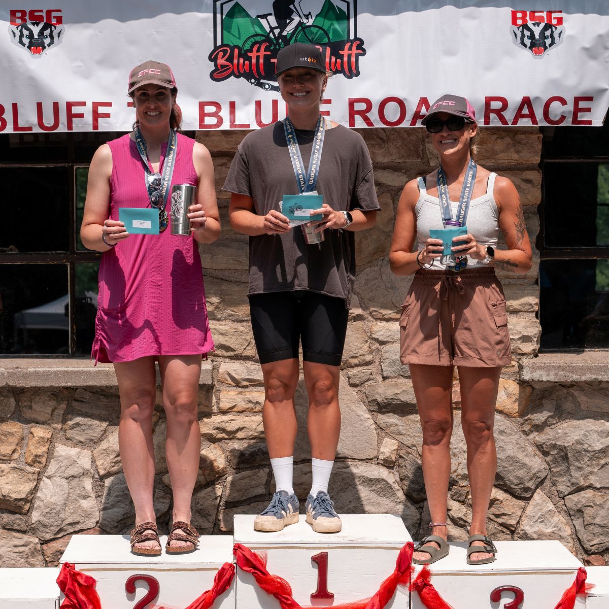 Three women stand on a white podium at the Bluff to Bluff Race, holding blue award certificates. A stone wall and race banner are visible in the background.