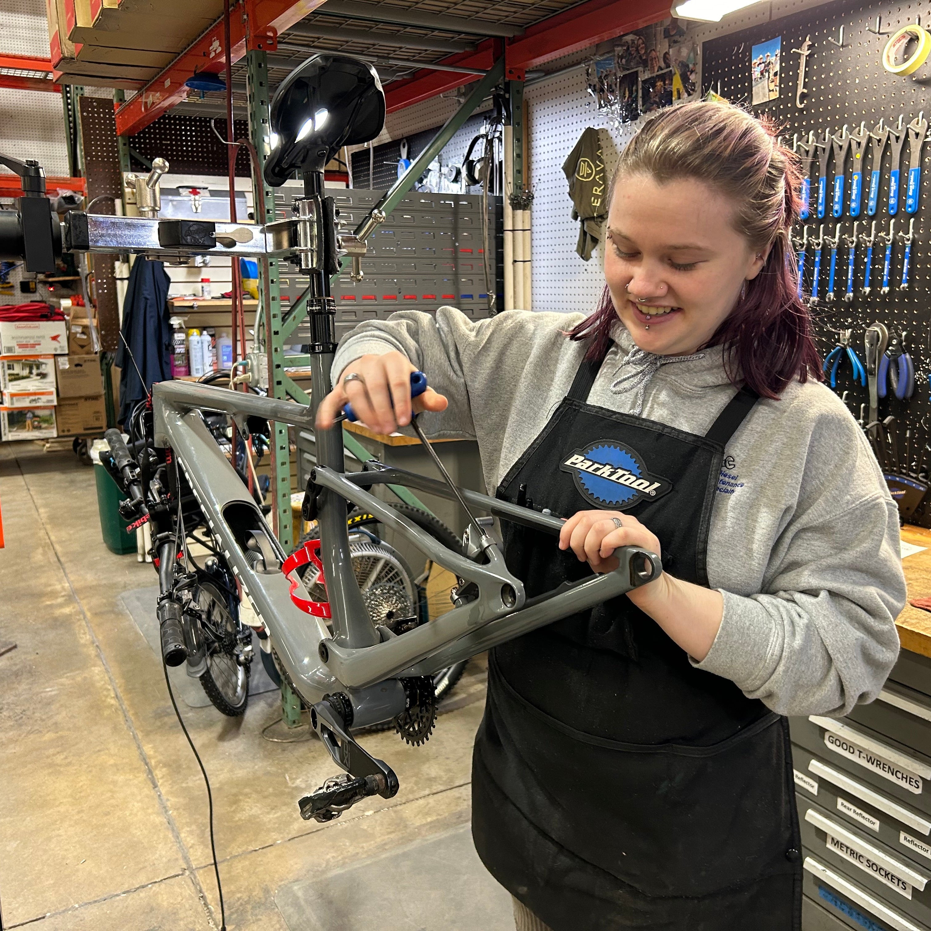 Hostel Shoppe mechanic Ava is holding a wrench to a bike frame in the service department's workshop.