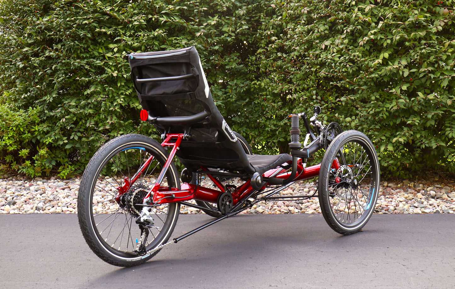 Rear angled view of the HP Velotechnik Gekko fx 20 Sport Recumbent Trike in Magma  on a bike path against a green leafy background.
