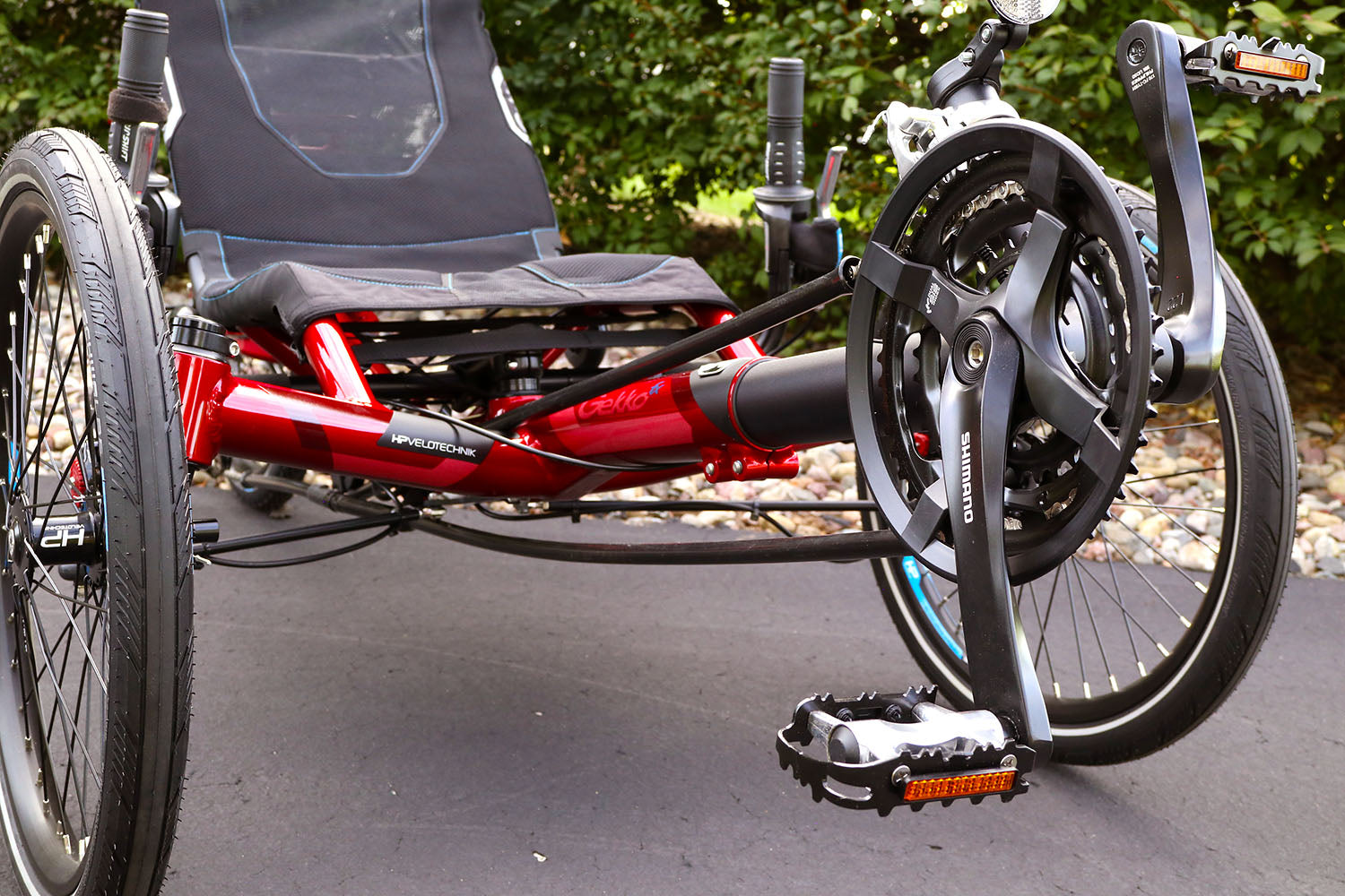 Closeup front view of the HP Velotechnik Gekko fx 20 Sport Recumbent Trike in Magma on a bike path against a green leafy background.