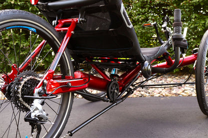 Closeup rear view of the HP Velotechnik Gekko fx 20 Sport Recumbent Trike in Magma on a bike path against a green leafy background.