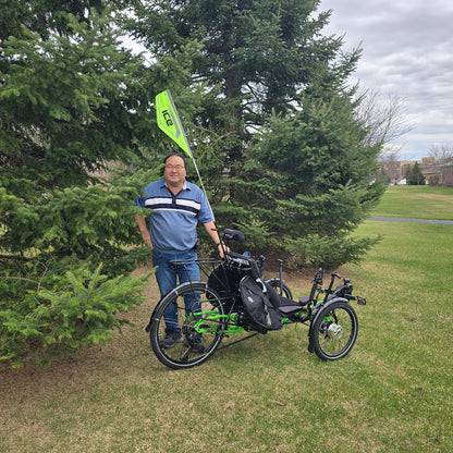 An ICE reflective safety flag mounted on an electric ICE trike on a rear light mount, sits next to a person for a height comparison. The flag is taller than the person, who is over 5 feet.