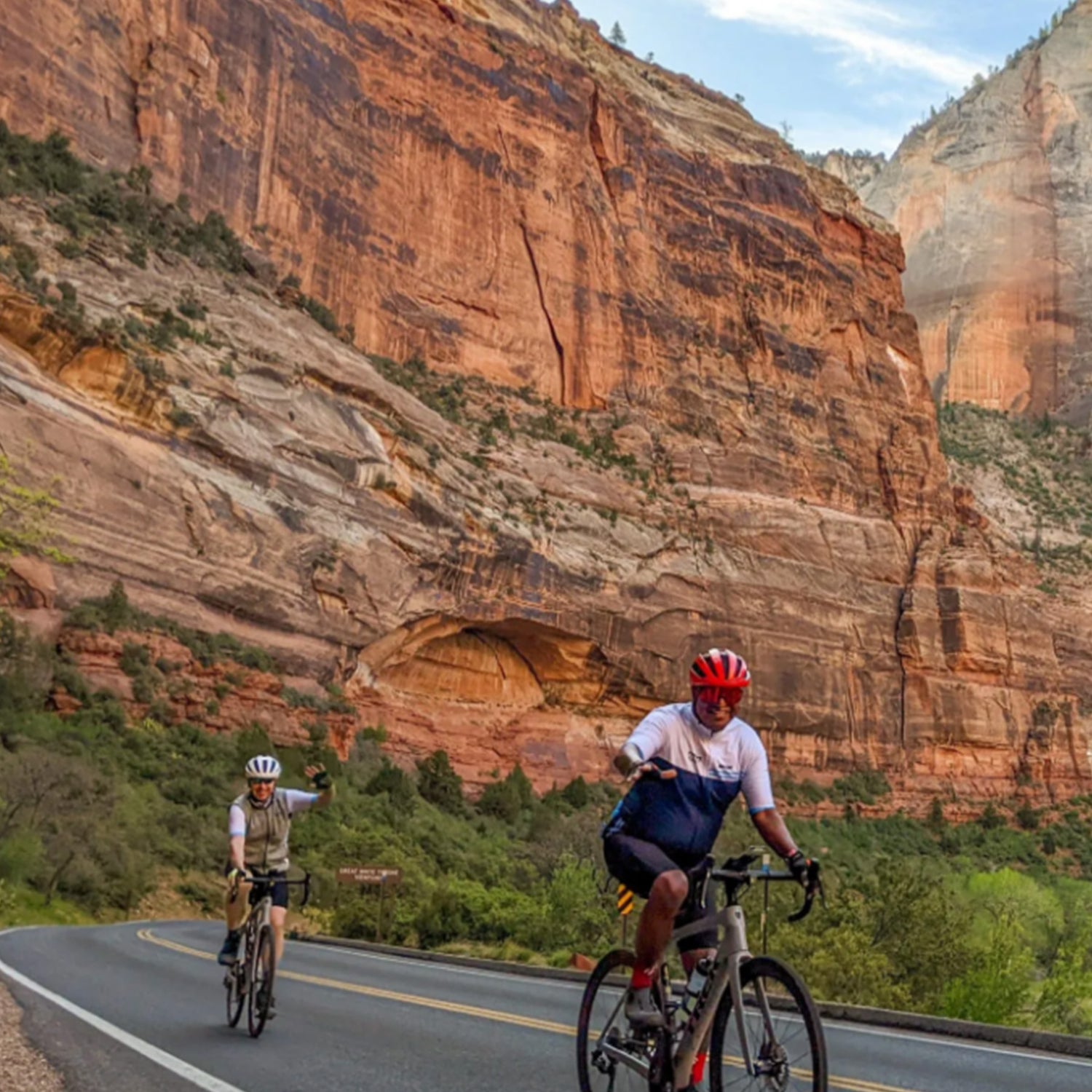 Two cyclists on a mountain road with red rock cliffs in the background.