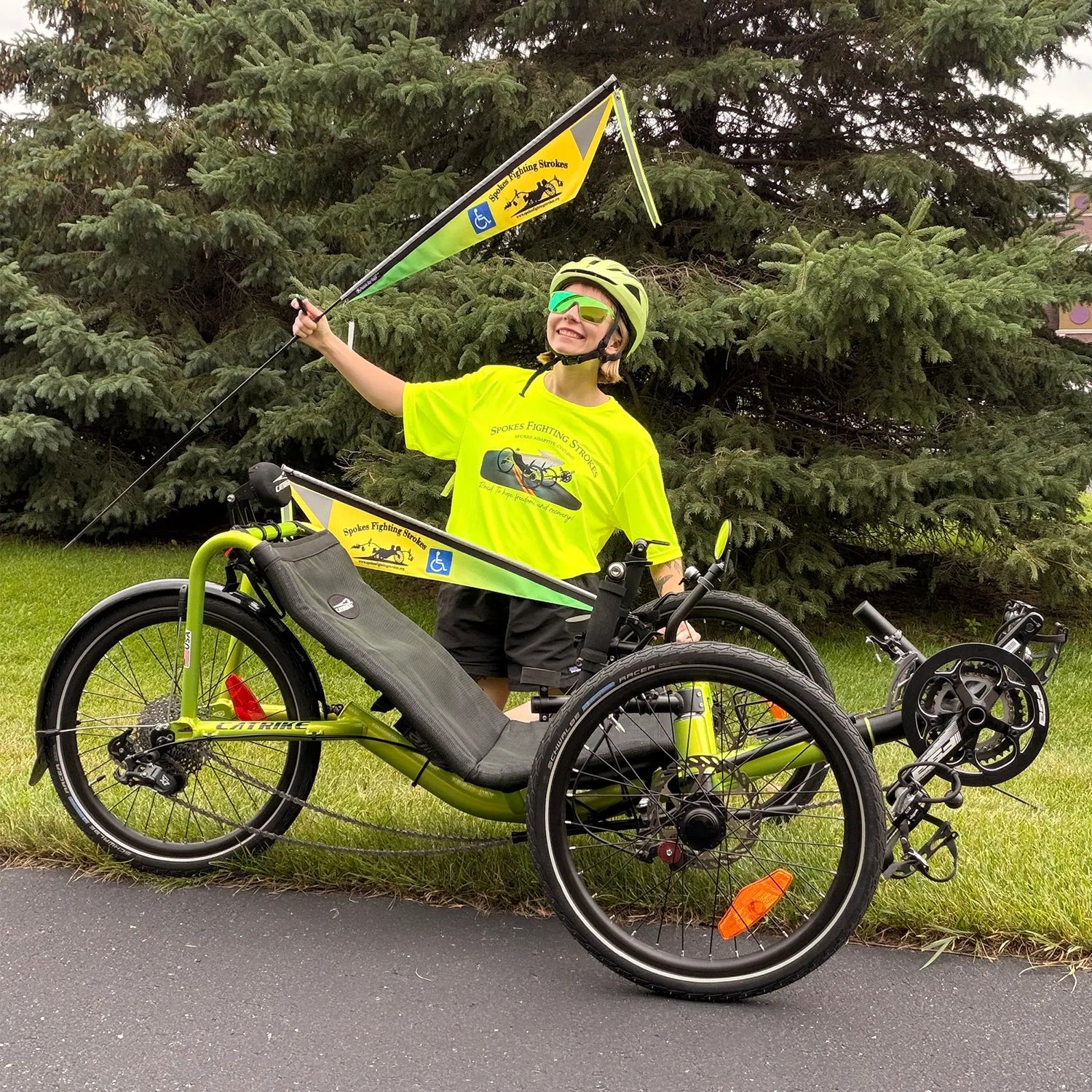 A person wearing a bright neon Spokes Fighting Strokes t-shirt, matching helmet and green sunglasses sits on a lime green recumbent trike with evergreen trees in the background. The trike has a matching yellow-green Spokes Fighting Strokes flag.
