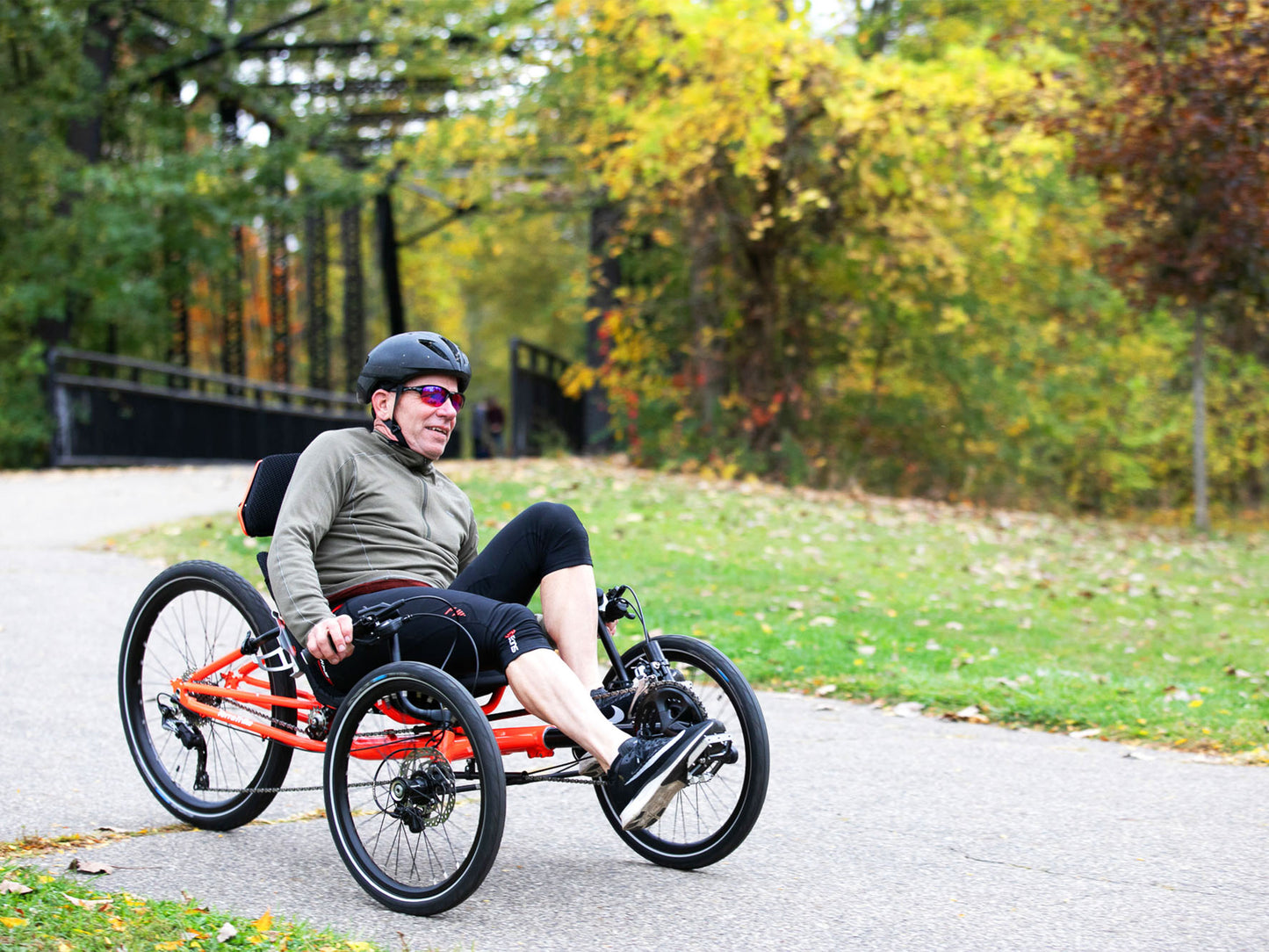 A man rides a TerraTrike GTS Recumbent Trike in Molten along a wooded bike path during fall.