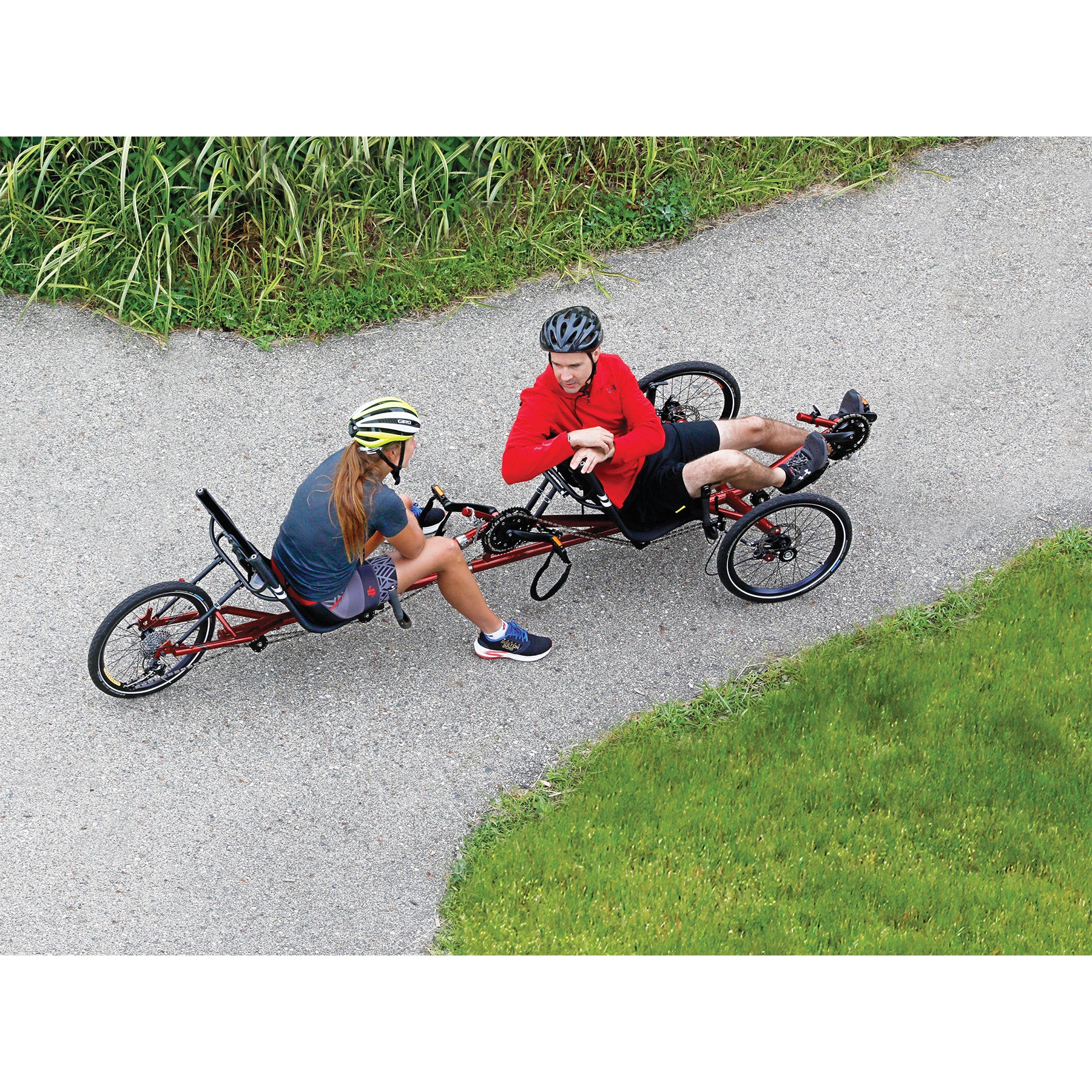 Two riders sit and converse on the TerraTrike Tandem Pro Sparkle Copper Recumbent Trike on a bike path surrounded by grass.