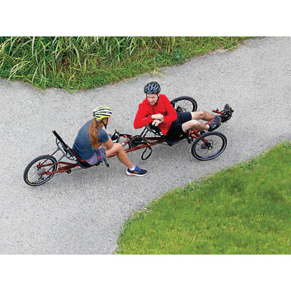 Two riders sit and converse on the TerraTrike Tandem Pro Sparkle Copper Recumbent Trike on a bike path surrounded by grass.