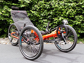 Three-wheeled orange recumbent trike on a paved path with greenery in the background.