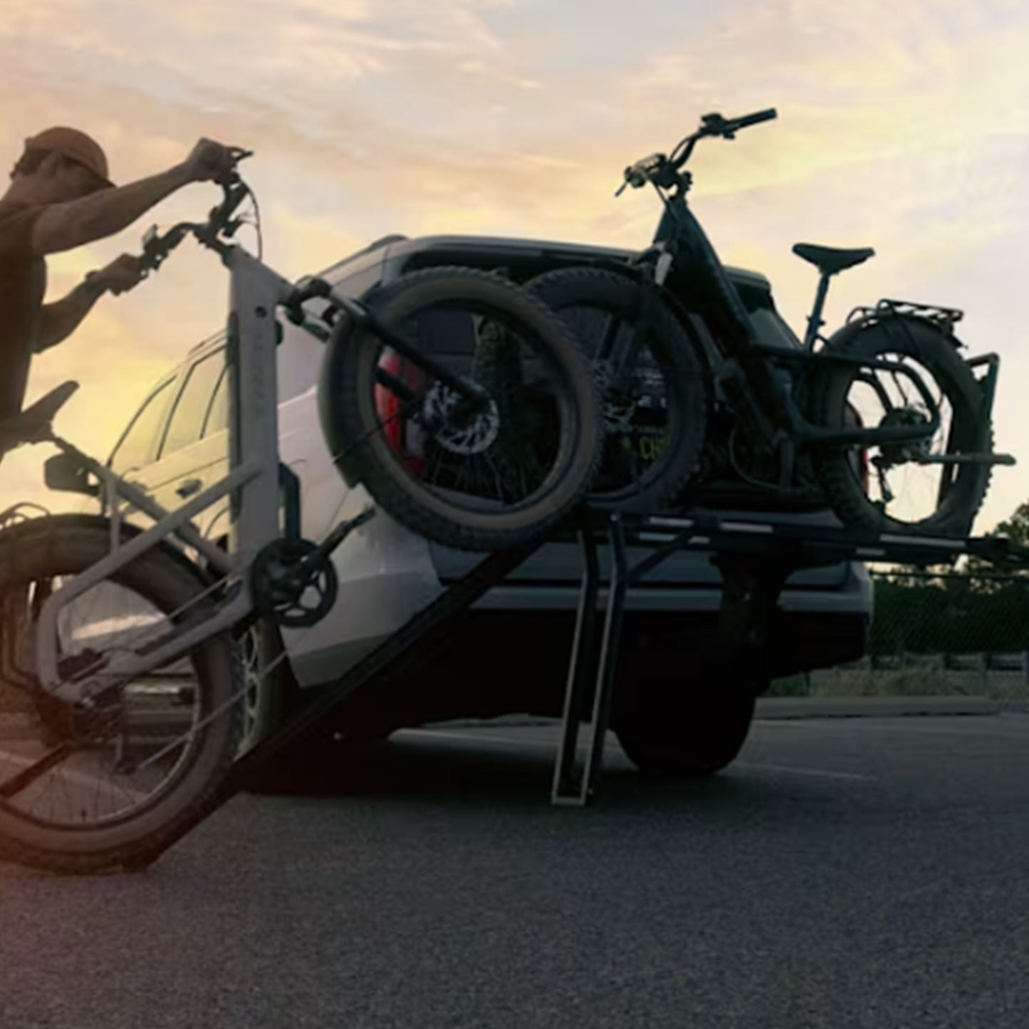 View of a man putting a Trek Borrego+ S on a car hitch rack which shows a second Borrego+ on the rack in an outdoor setting.
